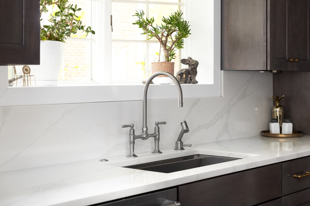 A kitchen faucet detail in a cozy kitchen with a stainless faucet, white marble countertop and backsplash, a bright window, and dark wood cabinets.