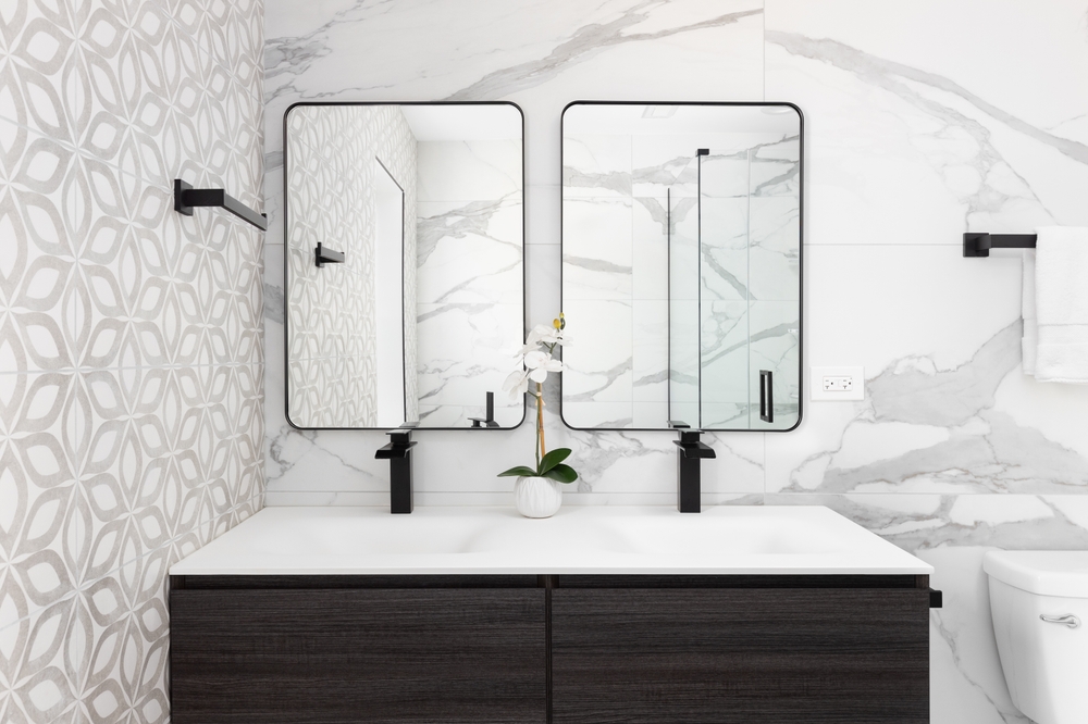 A bathroom detail with a modern wood cabinet, pattern and marble tiled walls, a white sink and black faucets, and black framed mirrors.