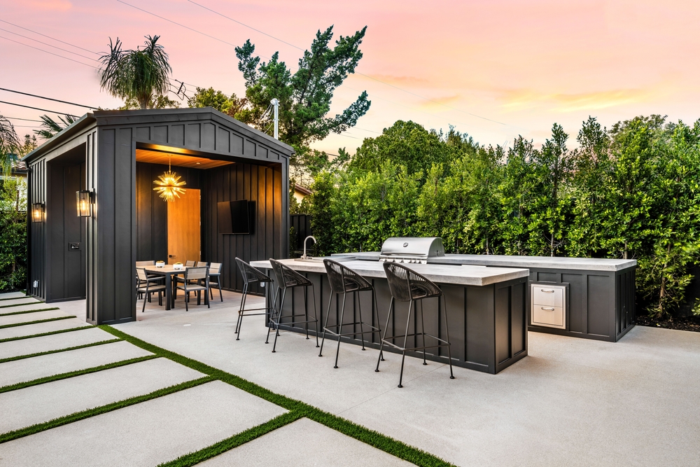 Modern outdoor kitchen and dining area with sleek black furniture and a stylish pergola, set against a lush green garden at sunset.