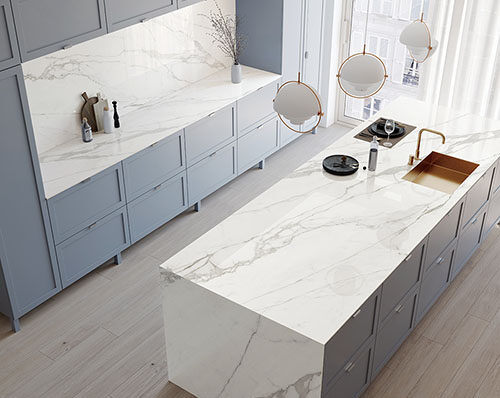 A modern white kitchen with porcelain countertops, backsplash, and island with light shining through the window. 