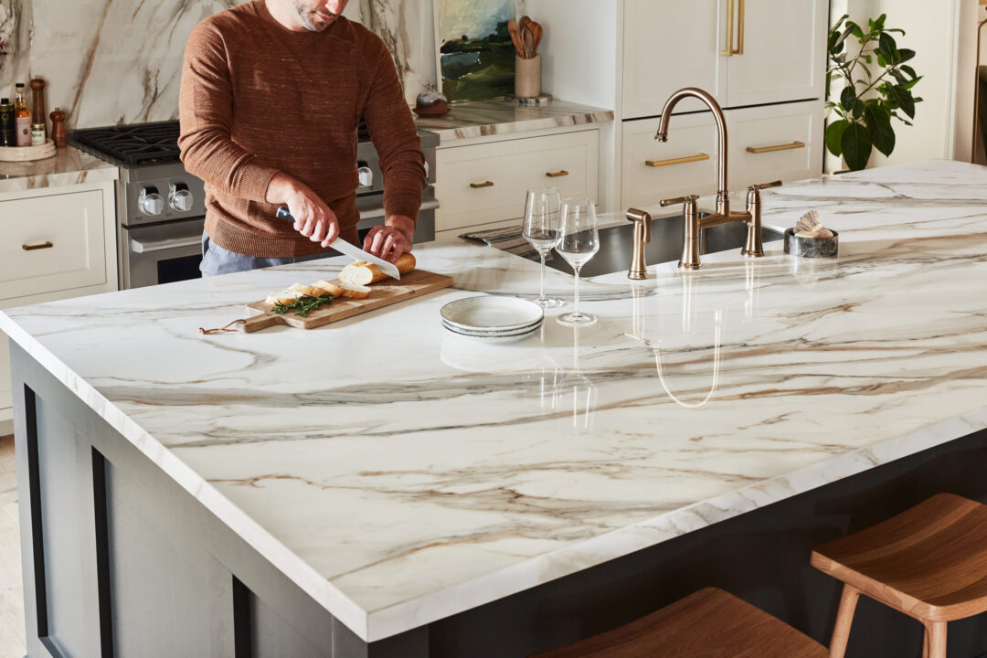 A man in a luxurious kitchen with upscale kitchen countertops slicing bread on a wood cutting board atop a white and gold porcelain surface.